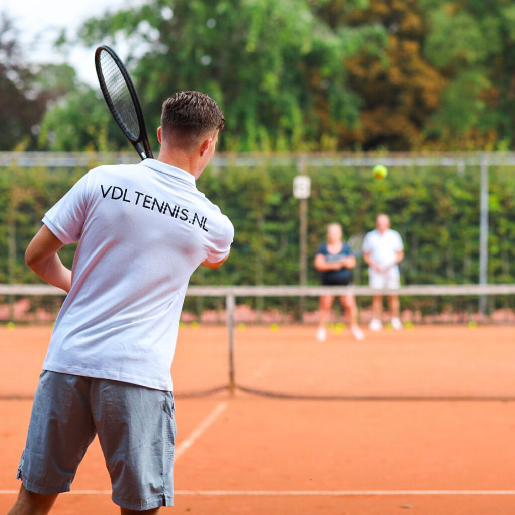 Tennisleraar met een lichtblauw t-shirt met vdltennis.nl op zijn rug slaat een tennisbal naar de andere kant van het net op een bruine tennisbaan.