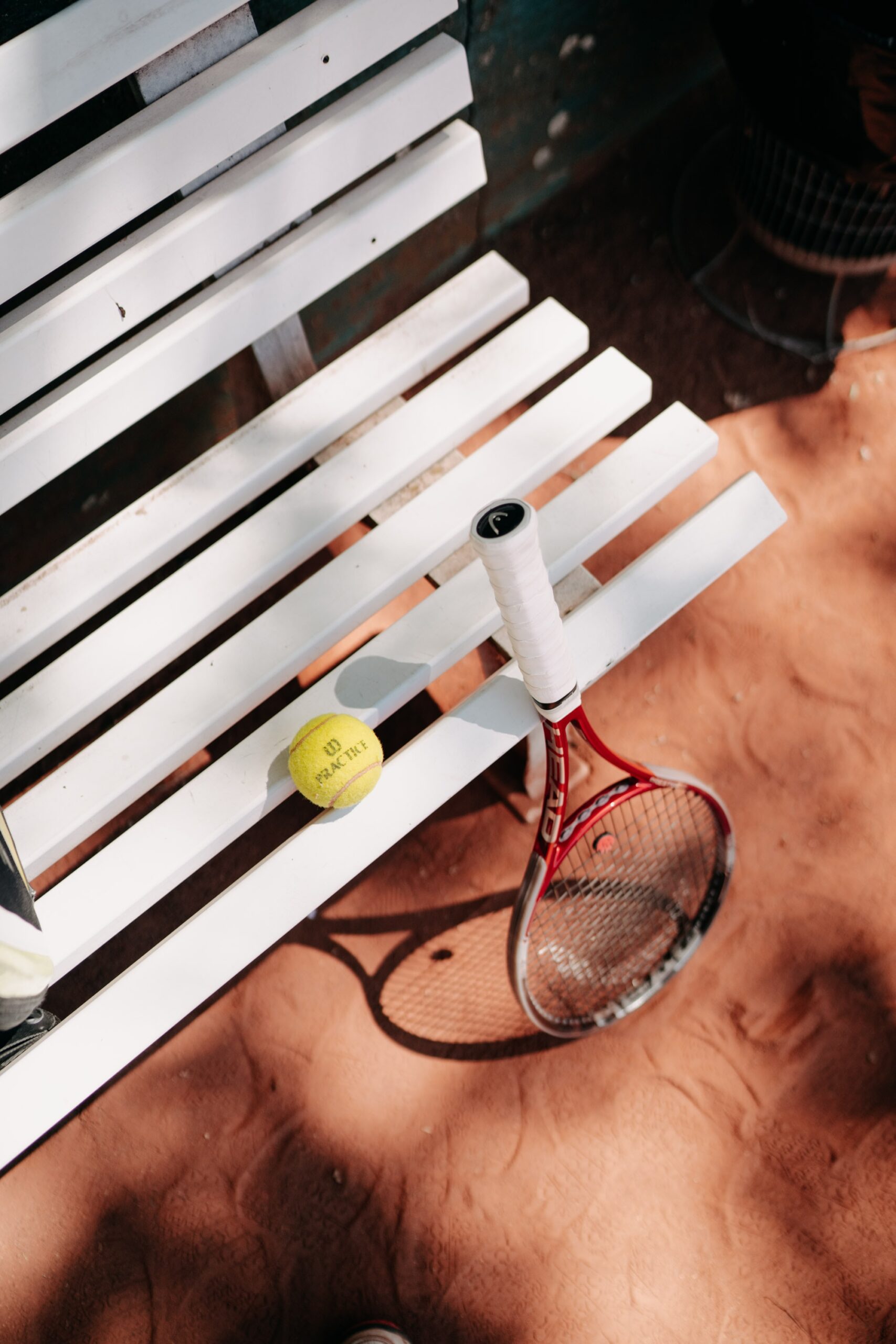 Tennisracket leunt tegen een wit bankje op de tennisbaan. Op het witte bankje ligt een lichtgroene tennisbal.