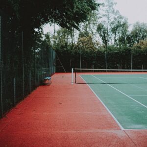 Groene tennisbaan met net in het midden. Op de achtergrond staan bomen. Tennisles in Schiedam worden op dit soort banen gegeven.