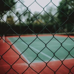 Tennisbaan gefotografeerd door een hek heen. tennisbaan is groen. Op de achtergrond staan bomen. Tennisles in Schiedam worden op dit soort banen gegeven.