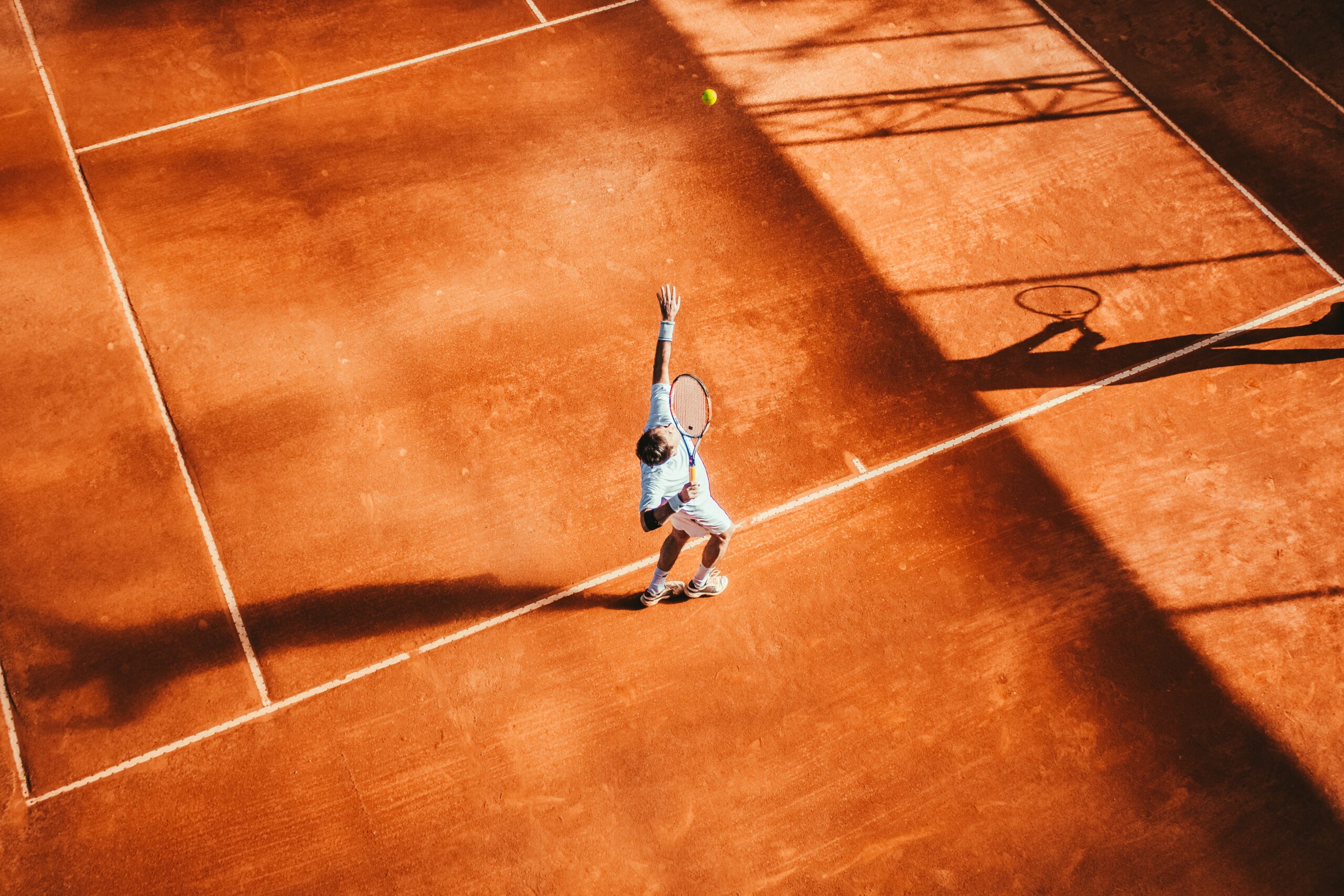Bruine tennisbaan. Iemand op de tennisbaan serveert een tennisbal. Tennisracket is in de lucht. Tennisbaan in Schiedam.