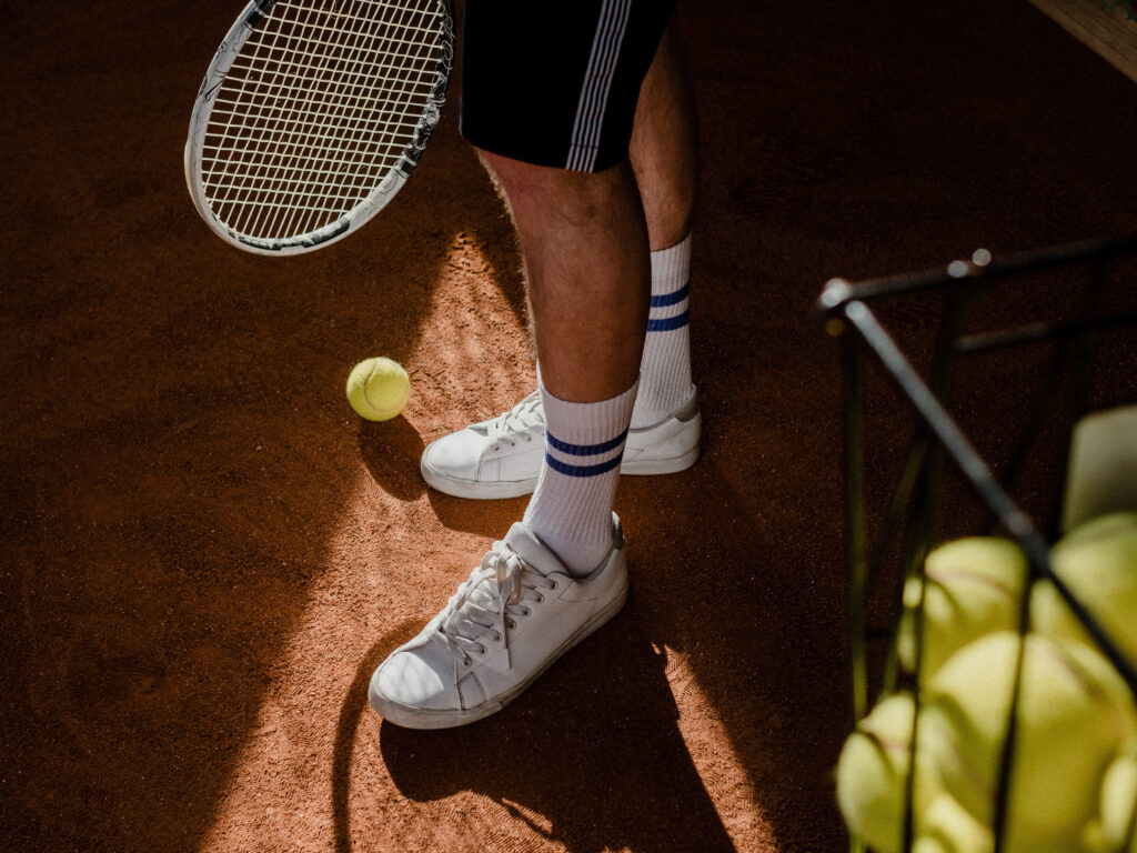 Tennisser staat op bruine tennisbaan met tennisracket in zijn hand. Naast hem staat een mandje met tennisballen er in.